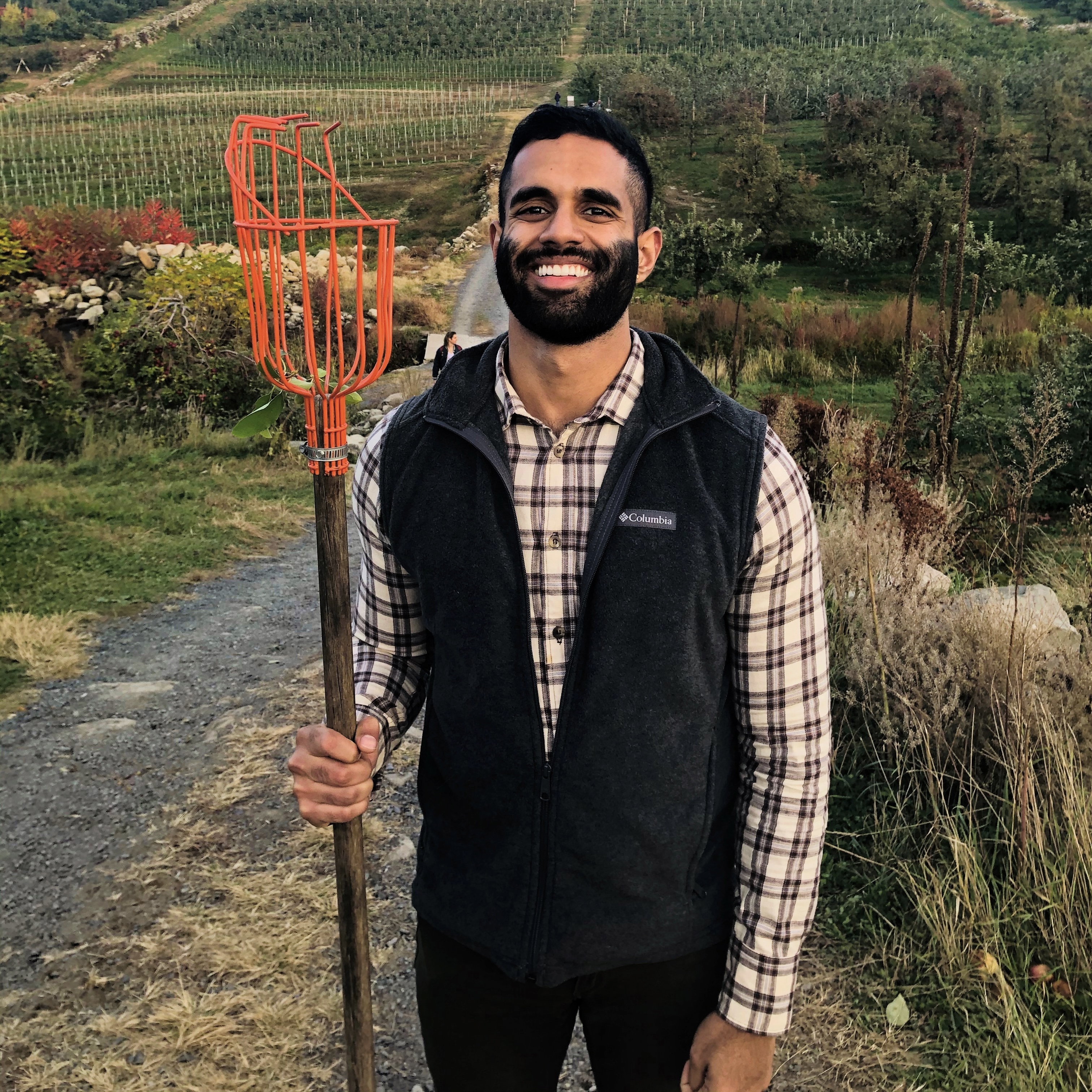 Shawn standing on a hiking trail wearing a plaid shirt and black Columbia vest, holding an orange rake